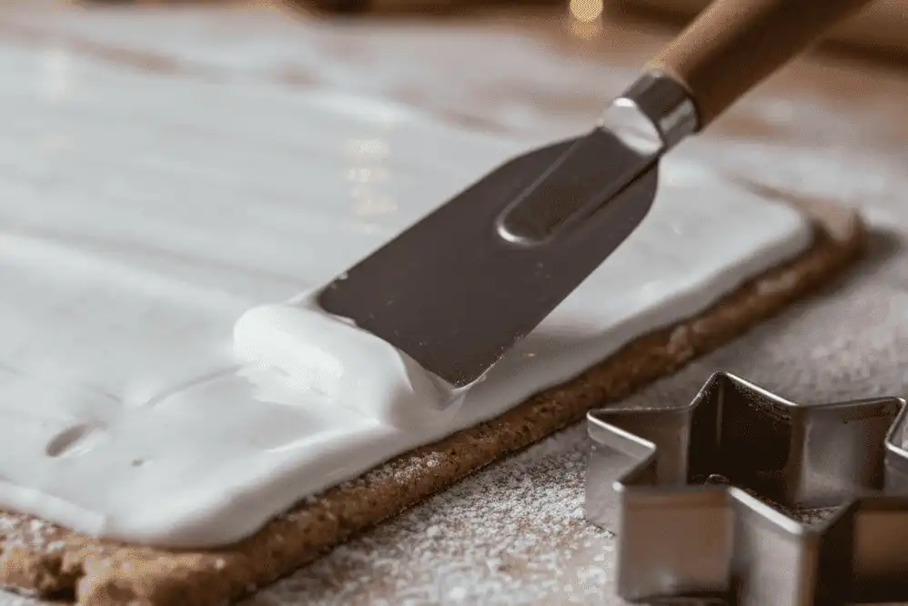 Spreading stiff white meringue glaze across rolled almond cinnamon dough using an offset spatula before cutting stars